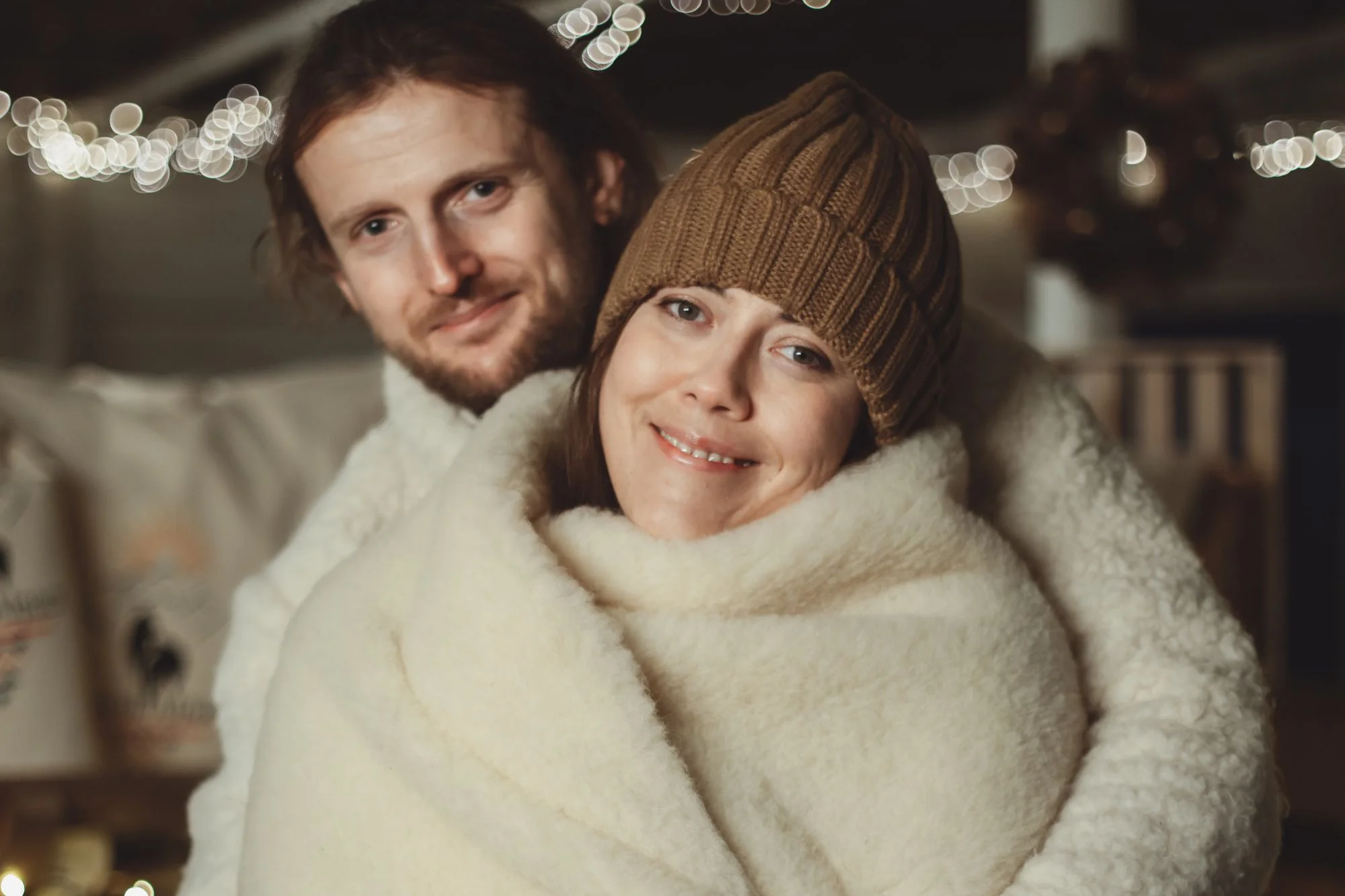 dwie osoby owinięte w ecru koc, mężczyzna z brodą i kobieta w brązowej czapce, w tle rozmyte światełka
