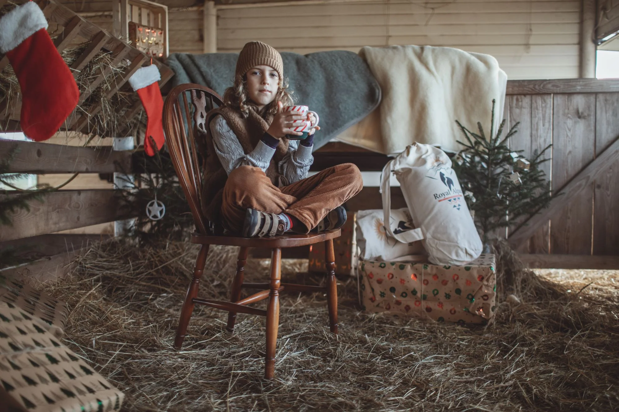 Chłopiec siedzący na krzesęłku w szpce. Ubrany w wełnianą kamizelkę. Trzyma kubek herbaty