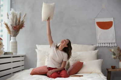 Uśmiechnięta kobieta siedzi na łóżku pościelonym kołrą i poduszkami z serii merino line. Podnosi poduszkę ponad głowę.