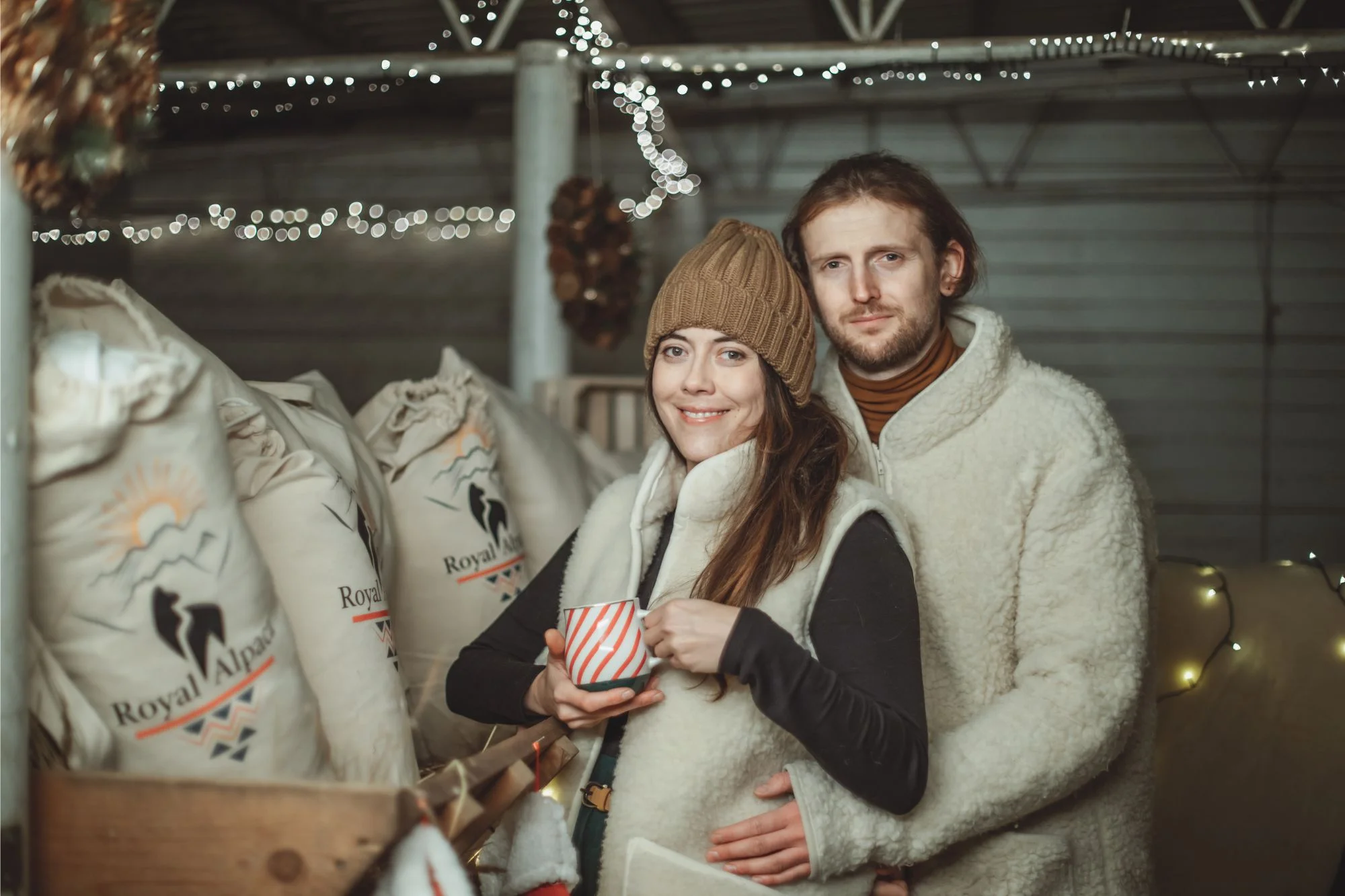 Para w wełnianych kamizelkach. Mężczyzna obejmuje kobietę. Kobita trzyma kubek z gorącym napojem. W tle worki z produktami Royal Alpaca.