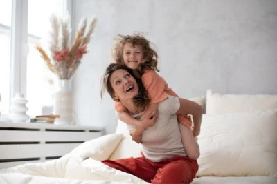 Kobieta siedzi w łóżku z chłopcem na plecach. Na łóżku kołdra i poduszki z wełny merino