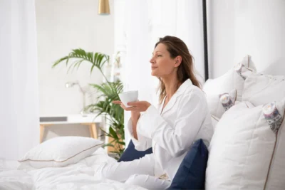 Kobieta w białej piżamie, siedzi w łóżku i popija kawę