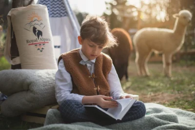 Chłopiec w wełnianej kamizelce, siedzi na trawie na kocu. Za nim alpaki
