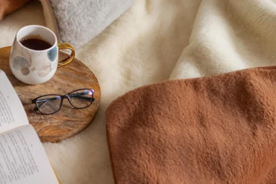 Zbliżenie na łóżko na którym leży kołdra i poduszka wełniana, książka oraz kubek z kawą i okulary