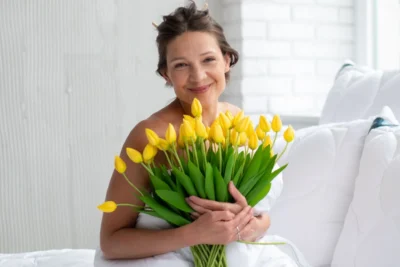 Kobieta zawinięta w białą kołdrę, trzyma w rękach duży bukiet żółtych tulipanów.