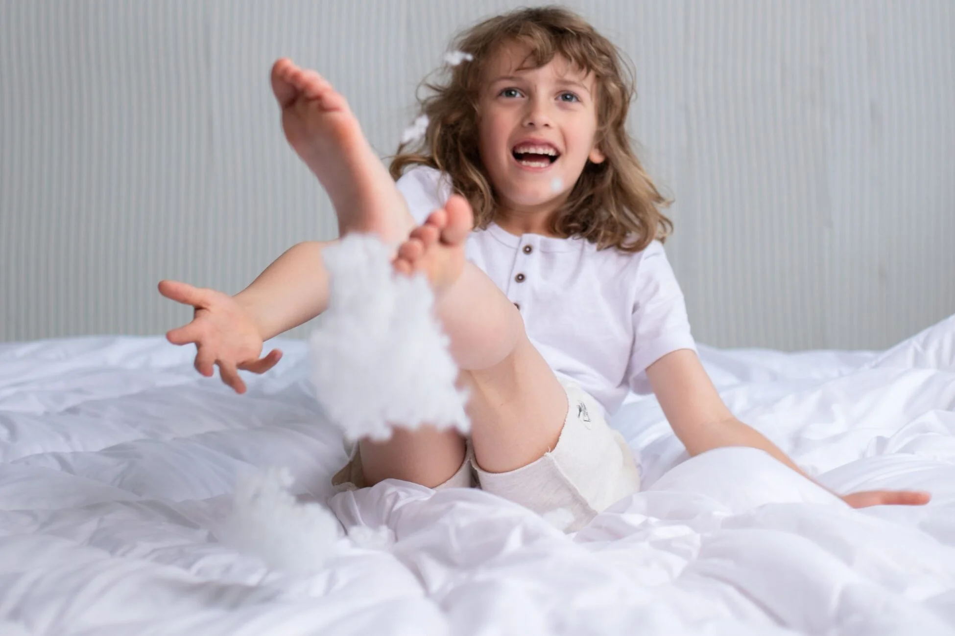 Uśmiechnięty chłopiec skacze po łóżku i rzuca wypełnieniem poduszki