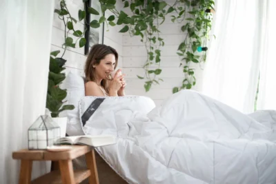 Kobieta leży w łóżku, pije herbatę. Pzzykryta biała kołdrą.