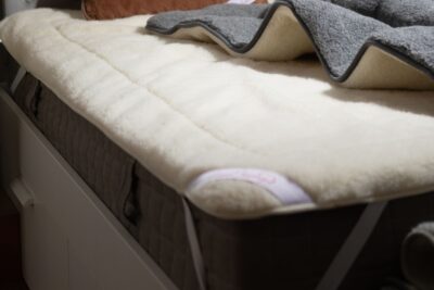 wełniany ochraniacz materac na łóżku z szarym kocem i brązową poduszką w tle