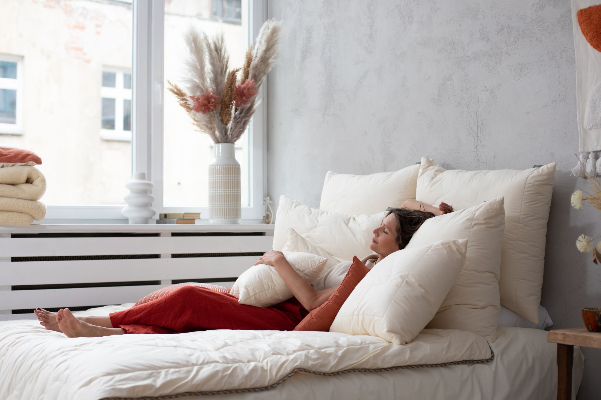 Kobieta leży na łóżku otoczona poduszkami, w tle okno i dekoracje wnętrza