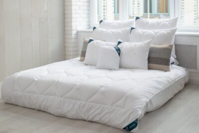 Łóżko, na nim biała kołdra i liczne poduszki. Na łóżku podkład na materac pod prześcieradło