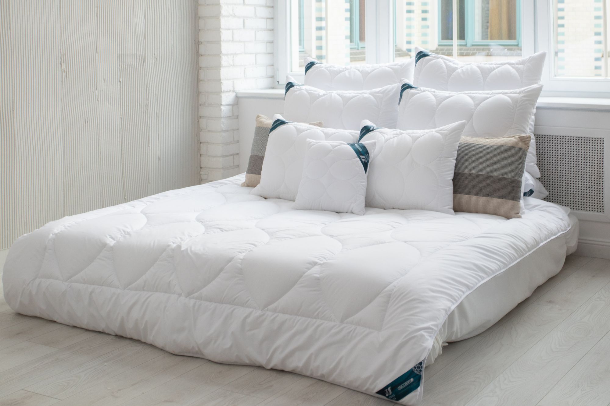 Łóżko, na nim biała kołdra i liczne poduszki. Na łóżku podkład na materac pod prześcieradło