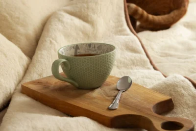 Zielony kubek z kawą, obok łyżeczka, leżą na drewnianej deseczce. Deseczka położona na wełnianą kołdrę.