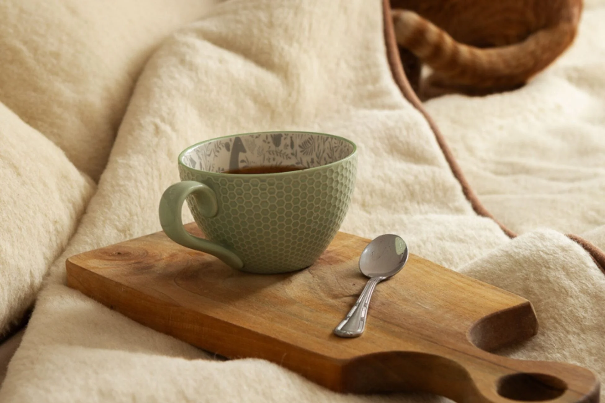 Zielony kubek z kawą, obok łyżeczka, leżą na drewnianej deseczce. Deseczka położona na wełnianą kołdrę.