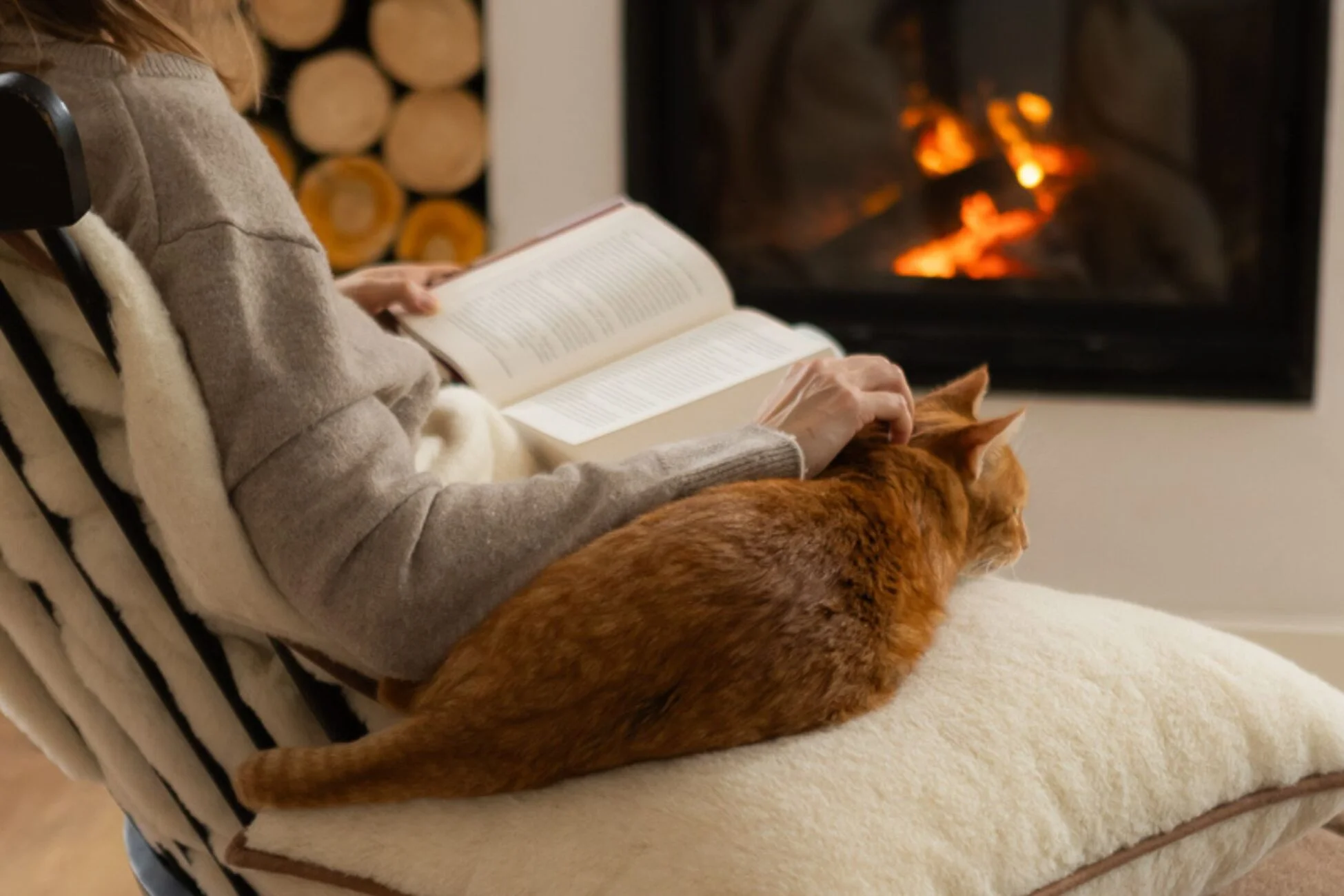 Kobieta siedząca na siedzisku z wełny, czyta książkę. Obok śpi rudy kot. W tle kominek.