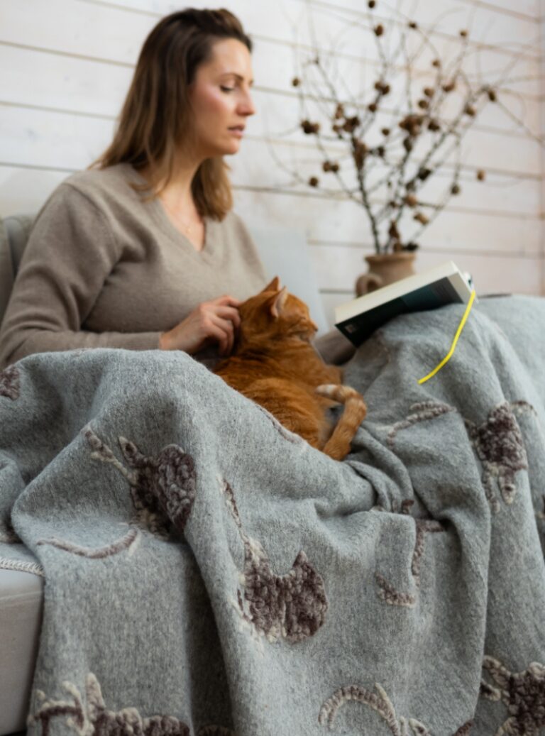 Kobieta siedząca na kanapie z książką i rudym kotem, przykryta szarym wełnianym kocem ze wzorem jeleni