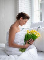 Kobieta owinięta w białą kołdrę trzyma bukiet żółtych tulipanów w jasnym wnętrzu