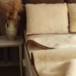 Poduszki w jasnym kolorze na łóżku z wełnianą kołdrą ecru w obok wazonu z roślinami na stoliku nocnym.