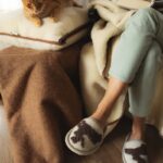 Osoba w szarym swetrze siedzi na kanapie, obok niej leży kot, a na kolanach koc i poduszka z wełny. Na stopach ma wełniane kapcie ze wzorem jelonka