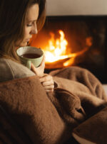 Kobieta siedząca przy kominku, trzymająca filiżankę, owinięta w brązowy koc