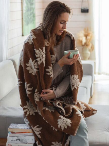 Kobieta siedząca na kanapie, owinięta brązowym kocem z białymi wzorami, trzyma filiżankę w dłoniach