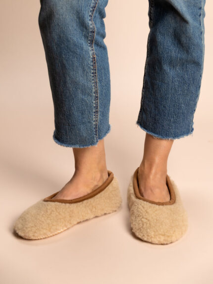 beige, geschlossene Wollhausschuhe an den Füßen, Jeans mit ausgefransten Hosenbeinen, neutraler Hintergrund