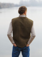 mężczyzna w ciemnozielonej kamizelce wełnianej stoi plecami nad wodą w mglisty dzień