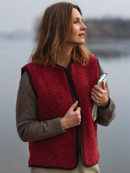 Kobieta w czerwonym bezrękawniku trzyma książkę, stoi nad wodą w mglistym otoczeniu