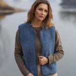 Kobieta w niebieskiej kamizelce i brązowym swetrze stoi nad wodą w otoczeniu natury