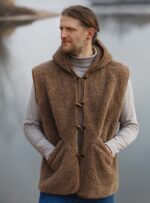 Mężczyzna w brązowej kamizelce z kapturem stoi nad wodą w naturalnym otoczeniu