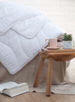 Biała kołdra na łóżku, obok stoliczek z filiżanką i książką na podłodze