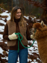 Kobieta w brązowej kamizelce i rękawiczkach trzyma linkę prowadząca alpakę w zaśnieżonym lesie