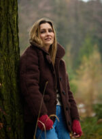 Kobieta w ciemnobrązowej kurtce wełnianej opiera się o drzewo w lesie, nosi czerwone rękawiczki