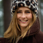 Kobieta z uśmiechem w futrzanej opasce z wzorem panterki, w brązowym płaszczu wełnianym w lesie