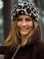 Kobieta z uśmiechem w futrzanej opasce z wzorem panterki, w brązowym płaszczu wełnianym w lesie