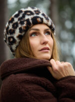 Kobieta w brązowej kurtce z wełny w opasce w wzór panterki, patrzy w górę na tle lasu