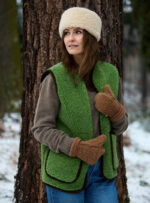 Kobieta w zielonej kamizelce z wełny, brązowych rękawiczkach i jasnej opasce na głowie, stoi przy drzewie w lesie