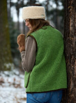 Kobieta w zielonej kamizelce i brązowych rękawiczkach stoi tyłem przy drzewie w lesie