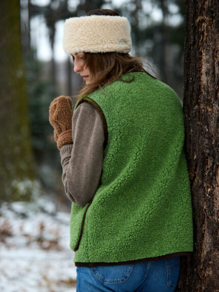 Kobieta w zielonej kamizelce i brązowych rękawiczkach stoi tyłem przy drzewie w lesie