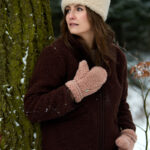 Kobieta w bordowej kurtce wełnianej i beżowych rękawicach stoi przy drzewie w lesie. Zimowa aura.