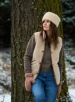 Kobieta w beżowej kamizelce i czapce, oparta o drzewo w zimowym lesie