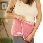 Kobieta w jasnym topie zakłada różowy pas z wełny, w tle widać zieloną kanapę i kota