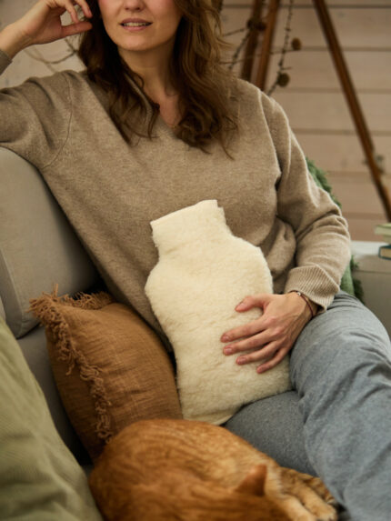 Kobieta w beżowym swetrze trzyma termofor w jasnym pokrowcu z wełny, obok leży kot i poduszki dekoracyjne