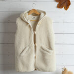 Jasna kamizelka z wełny alpaki z kapturem na drewnianym wieszaku na tle białej ściany.