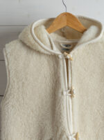 Jasna kamizelka z wełny alpaki z kapturem na drewnianym wieszaku na tle białej ściany.