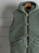 Szałwiowa kamizelka damska z kieszenią i zapięciem na sznurku na tle jasnych desek.