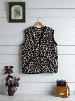 Kamizelka w wzór panterki na wieszaku, obok doniczka z rośliną i aparat fotograficzny na stole