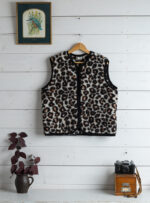 Kamizelka wełniana w wzór panterki na wieszaku, obok doniczka z rośliną i stary aparat
