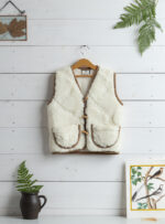 wełniana kamizelka w kolorze ecru z brązowymi detalami na wieszaku, obok roślina w doniczce