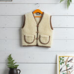 kamizelka ecru z wełny na wieszaku z brązowymi detalami, w tle białe deski