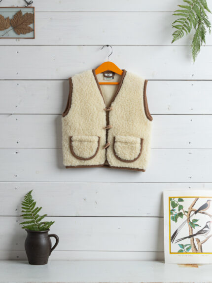 kamizelka ecru z wełny na wieszaku z brązowymi detalami, w tle białe deski